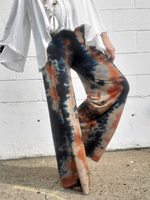 Person wearing tie-dye pants with a white shirt against a white brick wall.