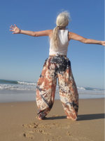 Person standing on a beach with arms outstretched, wearing a white top and patterned pants. Puffy harem pants in linen-raimie
