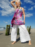 Woman in colorful top and white pants standing on a beach with blue sky and clouds.