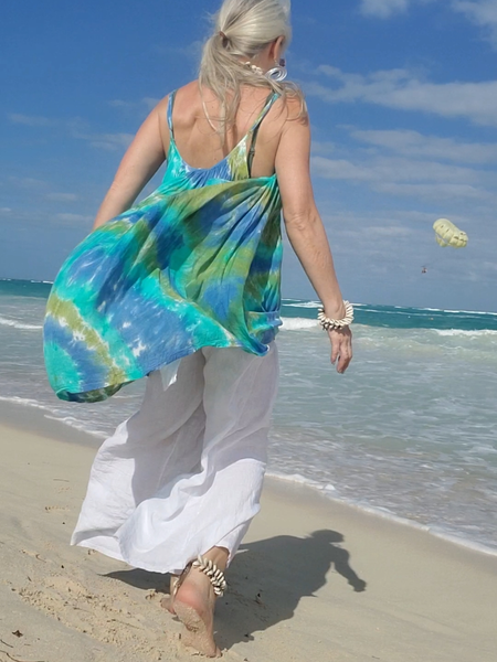 A mostly back view of me walking on the beach; the adjustable strap one size rayon parachut dress is floating behind me in the wind. hand dyed in aqua-blue-avocado colors. I'm wearing a pair of wide leg linen pants under