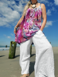 Woman in colorful top and white pants standing on a beach with blue sky and clouds.