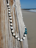 A beaded necklace draped over a weathered wooden post at the beach, featuring white baroque pearls and dark spacer beads, with a section of blue-green beads on the right side. The background shows a sandy beach and ocean under a clear blue sky.