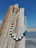 A beaded necklace with white baroque pearls and teal blue beads, draped over a weathered wooden post at a beach with ocean and sky in the background.