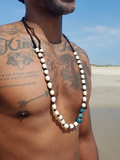 A man on a beach wearing a long beaded necklace made of mostly white beads with black accents and a few turquoise blue beads near the end.