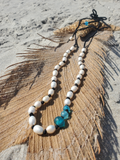 A beaded necklace with white round beads, black spacer beads, and three translucent blue faceted beads, displayed on a large brown feather over sand.