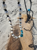 A beaded necklace with white pearl-like beads, black spacer beads, and turquoise blue accent beads, draped over a large tan feather on a sandy beach next to a starfish.
