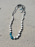 A beaded necklace with white round beads, black spacer beads, and two translucent blue faceted beads near the clasp, laid out on a sandy surface.