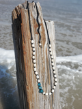 A beaded necklace with white round beads, black cord, and three blue faceted beads at the center, draped over a weathered wooden post with ocean waves in the background.