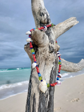 Colorful beaded necklace with shells draped over a piece of driftwood on a beach.