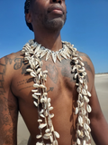 A man on a beach wearing a large necklace made of white cowrie shells and a matching shell collar, with multiple strands draped over his chest.