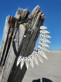handmade statement necklace made of white sliced shells with black braided cord, hanging from a weathered wooden post on a sandy beach under a clear blue sky.