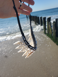 A handmade necklace featuring white seashells and black braided cord, held up by a hand against a beach and wooden pier background.