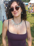 A woman with curly dark hair wearing round sunglasses, a dark plum camisole, and a chunky white and black statement necklace, photographed at an outdoor festival.