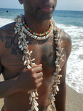 man wearing beaded jewelry at the beach. A cowrie shell choker with red green orange  and black and white beads, and 4 long necklaces made with hemp and cowrie shells