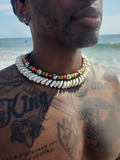 A close-up lifestyle shot of a man's neck and chest at the beach, wearing a double-strand shell necklace with white cowrie shells and a second beaded necklace with colorful round beads.
