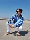 Man in a blue tie-dye shirt and white pants sitting on a beach.