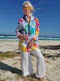 Woman in a colorful jacket and white pants standing on a beach with ocean waves in the background.