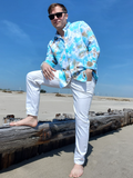 Man in a floral shirt and white pants sitting on a wooden log on a beach.