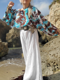 Person wearing a tie-dye shirt and white pants standing on a rocky beach.