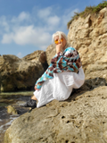 Person sitting on a rock by the ocean wearing a colorful jacket and white dress.