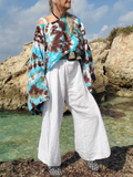 Person wearing a tie-dye shirt and white pants standing by a rocky coastline.