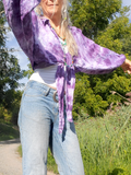 Person wearing a purple tie-dye shirt and light blue jeans standing outdoors with greenery in the background