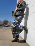 Person wearing tie-dye pants and a leather jacket leaning against a white wall.