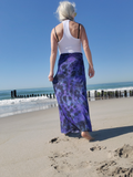 Woman walking on a beach wearing a white tank top and purple tie-dye skirt.