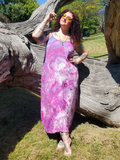 A woman wearing a sleeveless tie-dye maxi dress in shades of pink, purple, and white, standing outdoors in front of a large weathered tree trunk on a grassy lawn.