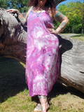 A woman wearing a pink and purple tie-dye maxi dress, leaning against a large fallen log in an outdoor park setting.