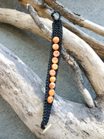 Black braided cord with orange beads on a piece of driftwood.