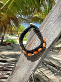 Black braided bracelet with orange beads on a wooden surface with palm leaves in the background