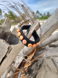 Black braided bracelet with orange beads on a piece of driftwood by the beach.