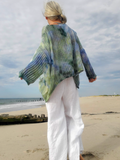 A woman standing on a sandy beach seen from behind, wearing a loose, long-sleeve ribbed knit top with a green, blue, and purple tie-dye pattern, paired with white pants.