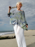 A woman standing on a sandy beach wearing a loose, oversized ribbed knit top with a blue, green, and gray watercolor-style pattern, paired with loose white pants.