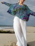 A woman on a sandy beach wearing a loose, long-sleeve knit top with a blue, green, and purple tie-dye pattern, paired with white wide-leg pants.