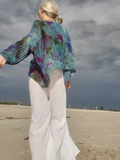 A woman standing on a sandy beach under a cloudy sky, wearing a loose, multicolored knit sweater with blue, purple, and green tones, paired with white wide-leg pants.