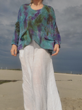 A person standing outdoors on a sandy beach wearing a loose, long-sleeve knit top with a teal, purple, and olive tie-dye pattern, paired with loose white pants.