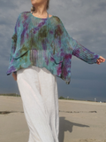 A woman standing on a sandy beach wearing a loose, long-sleeve knit top with a teal, purple, and olive tie-dye pattern, paired with white wide-leg pants.