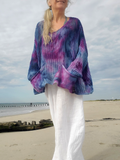 A woman standing on a sandy beach wearing a loose, long-sleeve knit top with a blue, purple, and pink tie-dye pattern, paired with white pants.