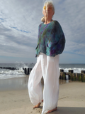 An older woman stands on a sandy beach wearing a loose, long-sleeve pleated knit top with a blue, green, and purple tie-dye pattern, paired with wide-leg white pants.