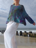 A woman standing on a beach wearing a loose, long-sleeve ribbed knit top with a blue, green, and purple tie-dye pattern, paired with white wide-leg pants.