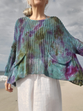 A woman wearing a loose, open-knit poncho-style sweater with a teal, green, purple, and blue tie-dye pattern, paired with white pants, standing on a sandy beach with the ocean and cloudy sky in the background.