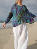 A woman standing on a sandy beach wearing a loose, open-knit long-sleeve top with a blue, purple, and green tie-dye pattern, paired with white pants.