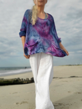 A woman standing on a sandy beach wearing a loose, oversized knit top with a blue, purple, and pink tie-dye pattern, paired with white wide-leg pants.