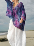 A woman standing on a beach wearing a loose, open-knit poncho-style top with a blue, purple, and pink tie-dye pattern, paired with white wide-leg pants.
