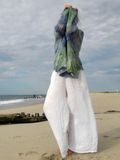 A person standing on a sandy beach with the ocean and cloudy sky in the background, wearing a green and blue striped knit top and loose white pants.