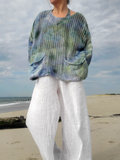 A person standing on a sandy beach wearing a loose, open-knit sweater with a blue, green, and gray tie-dye pattern, paired with white wide-leg pants.