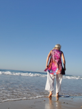 Person walking on a beach with a colorful top and white pants.