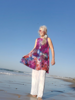 Woman in a colorful dress and white pants standing on a beach with blue sky.